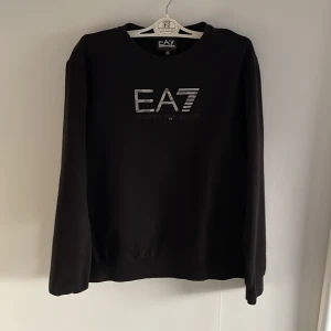 Svart tröja från EA7 - Säljer en stilren svart tröja från EA7 med deras logga tryckt på framsidan. Tröjan har långa ärmar och en klassisk passform. Perfekt för en avslappnad och trendig look.                                  Svarar snabbt på frågor!