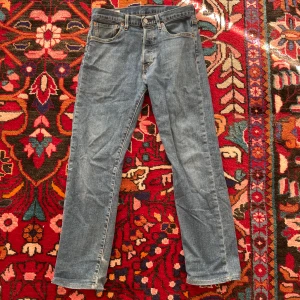 Levis 501 jeans - Klassiska Levis 501 jeans i blå denim. De har en rak passform och är tillverkade i slitstarkt bomullsmaterial. Jeansens design inkluderar fem fickor och en knappgylf. Perfekta för en tidlös stil.