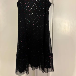 Svart prickig klänning - Snygg svart klänning med färgglada prickar och volangdetalj längst ner. Klänningen är ärmlös med smala axelband och har en luftig passform. Perfekt för en festlig kväll!