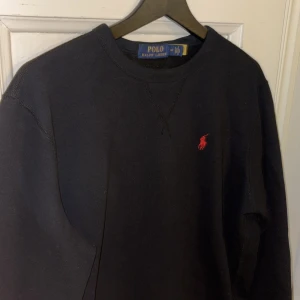 Svart tröja från Polo Ralph Lauren - Säljer en stilren svart tröja från Polo Ralph Lauren med det klassiska röda logotypbroderiet på bröstet. Tröjan har en rund halsringning och långa ärmar, perfekt för en avslappnad look.