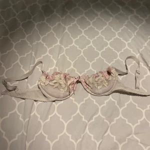 Vit och rosa bh med spetsdetaljer - Elegant bh i vitt med vackra rosa och vita spetsdetaljer. Den har justerbara axelband och en klassisk hakknäppning bak. Perfekt för att ge en romantisk touch till din underklädeskollektion.