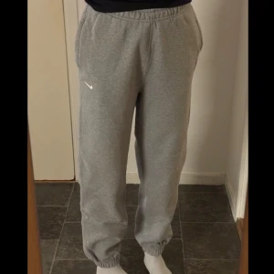 Grå fleecebyxor från Nike - Säljer ett par grå fleecebyxor från Nike med elastisk midja, snörning och fickor i sidorna. Byxorna har en avslappnad passform och är perfekta för en bekväm stil. Den klassiska Nike-loggan syns på sidan. Använd fåtal gånger