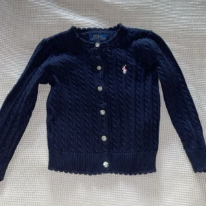 Mörkblå kofta från Ralph Lauren - Säljer en stilren mörkblå kofta från Ralph Lauren med kabelstickat mönster och knappar framtill. Den har långa ärmar och en liten logga på bröstet. Perfekt för att hålla sig varm och snygg under kyligare dagar. Svårt att hitta storlek mer än 2/2T och vad det motsvarar. Saknar lapp inuti plagg då det är lillasysters gamla och hon valt att ta bort pga den kliades 