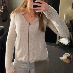 Beige kofta med dragkedja - Säljer en stilren beige kofta med dragkedja framtill. Koftan har en v-ringad design och långa ärmar, perfekt för en avslappnad look. Passar bra till både jeans och kjol.