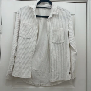 Vit randig skjorta - Säljer en stilren vit randig skjorta med långa ärmar och två bröstfickor. Skjortan har en klassisk krage och är tillverkad i ett mjukt material som ger en bekväm känsla. Perfekt för en avslappnad look.