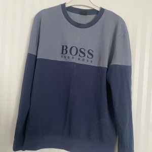 Blå och grå tröja från Hugo Boss - Snygg blå och grå tröja från Hugo Boss med stort BOSS-tryck på framsidan. Tröjan har långa ärmar och en bekväm passform. Perfekt för en stilren och avslappnad look. Den är som helt ny men jag säljer för att den inte passar med min stil längre. Pris går att diskuteras.