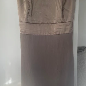 Beige långklänning - Elegant beige långklänning med en stilren design. Klänningen har en mjuk överdel och en plisserad nederdel som ger en fin silhuett. Perfekt för speciella tillfällen.