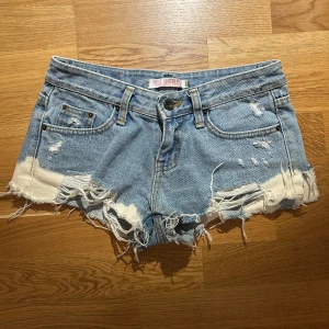 Blå jeansshorts från Blueray - Säljer ett par blå jeansshorts från Blueray med slitna detaljer och fransiga kanter. Shortsen har en lågmidjad passform och klassiska femficksdesign. Perfekta för en avslappnad stil.