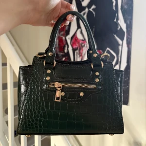 Grön handväska med krokodilmönster - Snygg svart handväska med krokodilmönster och guldfärgade detaljer. Väskan har en dragkedja framtill och två handtag för enkel bärning samt finns en långt band till också! 