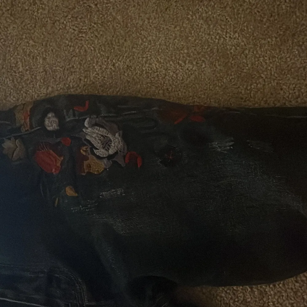 Snygga svarta bootcut jeans med färgglatt blommigt broderi på ena benet. Jeansen har en klassisk femficksdesign och är tillverkade i ett robust denimtyg. Perfekta för att ge din outfit en unik touch.. Farkut & Housut.