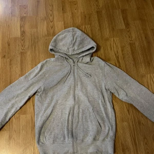 Grå hoodie med dragkedja - Säljer en stilren grå hoodie med dragkedja och huva. Tröjan har långa ärmar och praktiska fickor framtill. Perfekt för en avslappnad look.