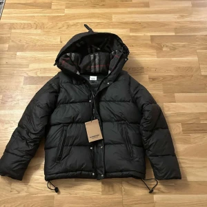 Svart pufferjacka från Burberry - Säljer en snygg svart pufferjacka från Burberry med huva och dragkedja. Jackan har ett klassiskt rutigt mönster på insidan av huvan och justerbara dragsnören. Perfekt för kyliga blir även väst pris kan diskuteras