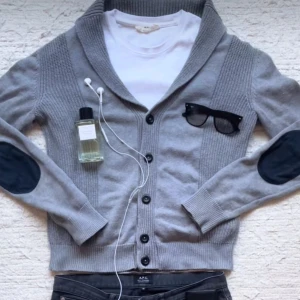 Grå kofta med knappar - Snygg grå kofta med knappar framtill och ribbade detaljer. Den har mörkblå armbågslappar som ger en stilren look. Storlek M men kan också passa S. Hör av dig vid frågor!