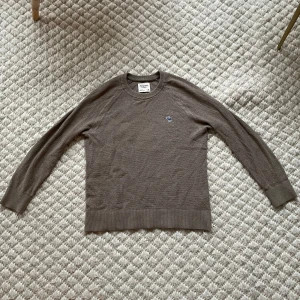 Mörk beige tröja från Abercrombie & fitch - Säljer en klassisk beige merinoulls tröja. Tröjan har långa ärmar och en liten broderad detalj på bröstet. Perfekt för en stilren och bekväm look. Använd fåtal gånger.