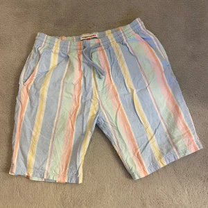 Randiga shorts från Tommy Jeans - Snygga randiga shorts från Tommy Jeans i pastellfärger som blå, gul, rosa och grön. De har en bekväm passform med snörning i midjan och sidofickor. Perfekta för en avslappnad sommarstil.