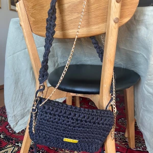 Svart virkad axelväska med kedjedetaljer - Snygg svart stickad axelväska med guldfärgade kedjedetaljer. Väskan har en praktisk dragkedja och en flätad axelrem för extra stil. Perfekt för att ge din outfit en unik touch.