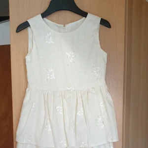 Champagnefärgad ärmlös klänning - Elegant champagnefärgad klänning med broderade blommor och volangdetaljer. Den är ärmlös med en dragkedja i ryggen och har en vacker, flerdelad kjol som ger en romantisk känsla. Barnstorlek ca 140