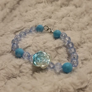 Blått armband med rosdetalj - Charmigt armband med ljusblå pärlor och en stor rosdetalj i mitten. Armbandet har en mix av genomskinliga och opaka pärlor som ger en fin kontrast. Perfekt för att lägga till en färgklick i din outfit.
