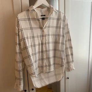 Vit och svart rutig skjorta från Lindex - Snygg vit skjorta från Lindex med svart rutmönster. Skjortan har långa ärmar och knappar framtill. Perfekt för en stilren look.