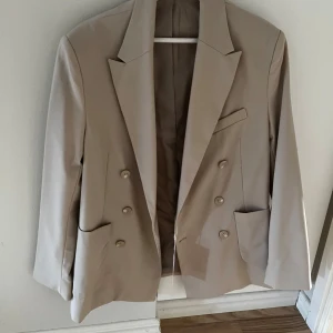 Beige kavaj från Bershka - Snygg beige kavaj från Bershka med dubbelknäppning och klassiska slag. Perfekt för en stilren look. Kavajen har två fickor framtill och en bröstficka för extra stilpoäng.