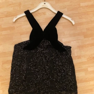Svart paljett-topp med rosett - Snygg svart topp täckt av glittriga paljetter. Den har en stor rosett vid halsen, vilket ger en elegant look. Perfekt för festliga tillfällen.