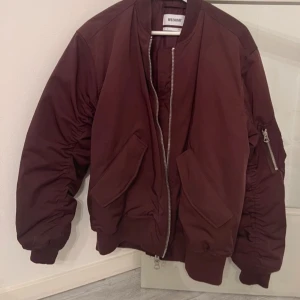 Mörkröd bomberjacka från Weekday - Snygg mörkröd klassisk bomberjacka från Weekday med dragkedja framtill och fickor på sidorna. Jackan har långa ärmar och är rätt oversized. Står storlek S men är något stor på mig som brukar bära storlek S vanligtvis. Lite tecken på användning men annars mycket fint skick!!