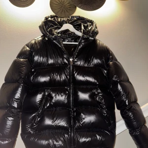 Svart pufferjacka från Zara - Säljer en snygg svart pufferjacka från Zara. Jackan har en glansig finish och är utrustad med dragkedja och huva. Perfekt för kyliga dagar när du vill hålla dig varm och stilren. 100% DUN