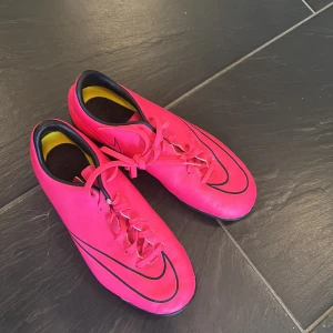 Rosa fotbollsskor från Nike - Snygga rosa fotbollsskor från Nike med svarta detaljer och snörning. Perfekta för inomhusspel med en slitstark sula. Skorna har en modern design och är lätta att bära.