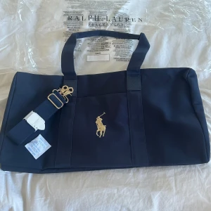 Blå väska från Ralph Lauren - Snabb köp=bra pris!! Snygg blå väska från Ralph Lauren med det ikoniska logotypen i guld. Väskan har två handtag och en avtagbar axelrem för extra bekvämlighet. Perfekt för att bära med sig det nödvändigaste med stil.