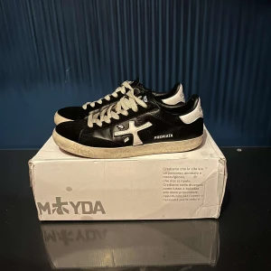 Premiata skor  - Säljer ett par snygga svarta sneakers från Premiata i mycket bra skick. De har vita detaljer och snörning, vilket ger dem en stilren look. Perfekta för både vardag och fest! Skostorlek 41.