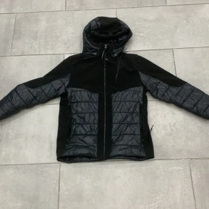 Svart och grå jacka från C.P. Company - Säljer en snygg svart och grå jacka från C.P. Company i 12 år. Jackan har en cool quiltad design med dragkedja och huva. Perfekt för höst och vinter, håller dig varm och stilren. Den är i mycket bra skick och redo för nya äventyr!