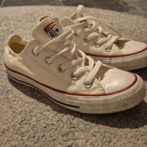 Vita Converse All Star sneakers - Säljer ett par klassiska vita Converse All Star sneakers i bra skick. De har den ikoniska röda och blå randen runt sulan och snörning framtill. Perfekta för en avslappnad stil och passar till det mesta i garderoben.