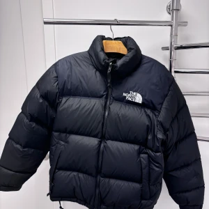 Svart pufferjacka från The North Face - Säljer en snygg och varm svart pufferjacka från The North Face. Perfekt för kalla vinterdagar med sin tjocka vaddering och höga krage. Den är i väldigt bra skick då den bara är använd i två vintrar. Köpt på junkyard. Svår att få tag på ny.