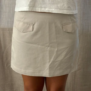 Beige kortkjol med fickdetaljer - Säljer en stilren beige kortkjol i mycket bra skick. Kjolen har två dekorativa fickor framtill!