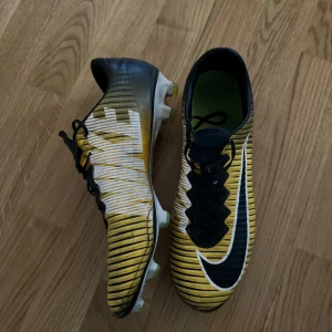 Nike Mercurial Vapor 11 - Säljer ett par coola fotbollsskor från Nike i gult och svart med snygga ränder. Skorna har snörning och är i bra skick, perfekta för fotbollsträningen. De har en grym passform och är designade för att ge bra grepp på planen.