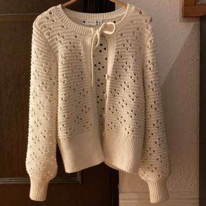 Vit stickad kofta med knytband - Superfin vit stickad kofta med ett sött knytband framtill. Den har ett vackert hålmönster och ribbade muddar vid ärmslut och nederkant. Perfekt för både vår och höst när du vill ha något mysigt men ändå stilrent. Passar till både jeans och klänning!