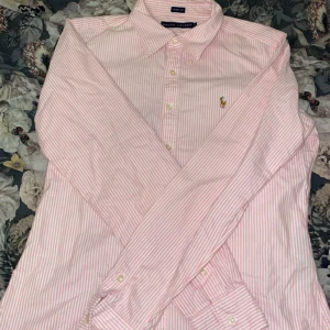 Rosa och vit randig skjorta från Ralph Lauren - Säljer en snygg rosa och vit randig skjorta från Ralph Lauren i skinny fit. Skjortan har långa ärmar och klassiska knappar framtill. Den är broderad med den ikoniska loggan på bröstet. Perfekt för både vardag och fest!