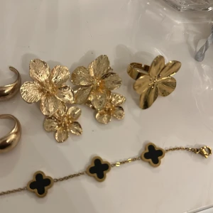 Guldiga smycken med blommotiv och svarta detaljer - Säljer ett set med guldiga smycken. Inkluderar stora blommiga örhängen, ett armband med svarta fyrklöverdetaljer, ett par guldiga hoops, en blom ring och 4 justerbara ringar. Perfekt för att piffa upp vilken outfit som helst! 🌟