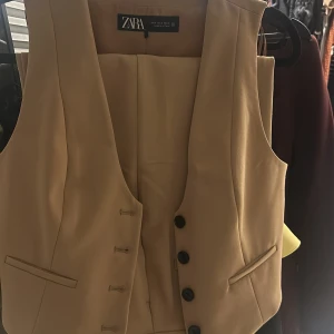 Beige väst och byxor från Zara - Snygg beige väst från Zara i ett stilrent och klassiskt snitt. Västen har knappar framtill och är perfekt för både vardag och fest. Den är i ett mjukt material som ger en bekväm passform. Passar bra till både byxor och kjol för en chic look.