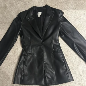 Svart skinnkavaj från H&M - Snygg svart skinnkavaj från H&M i storlek 34. Perfekt för en stilren look, med en knapp framtill och långa ärmar. Kavajen har en klassisk passform och är gjord i syntetiskt material som ser ut som skinn. Passar både till vardags och fest!