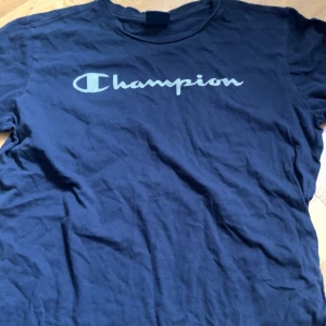 Mörkblå t-shirt från Champion - Säljer en mörkblå t-shirt från Champion i storlek 2X-Large men sitter som M. Den har en klassisk Champion-logga tryckt på bröstet. Perfekt för en avslappnad stil eller träning. T-shirten är i bra skick och gjord av bomull, vilket gör den bekväm att bära.