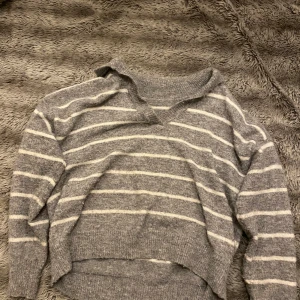 Grå randig stickad tröja - Säljer en supermysig grå stickad tröja med vita ränder. Den har en avslappnad passform och en snygg v-ringad krage. Perfekt för kyliga dagar när du vill vara både bekväm och stilren. Passar perfekt till jeans eller leggings!