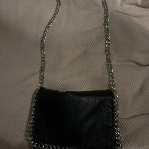 Tiamo väska  - Snygg svart axelväska i skinn med coola kedjedetaljer runt kanterna och som axelrem. Perfekt för en utekväll eller när du vill lägga till lite edge till din outfit. Väskan är i bra skick och har en stilren design som passar till det mesta.
