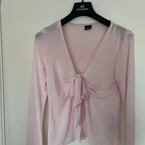 Ljusrosa långärmad topp från Ginatricot  - Säljer en söt ljusrosa långärmad topp från Gina. Den har en knytning framtill som ger en fin detalj och en v-ringad hals. Perfekt för vår och sommar, och kan enkelt kläs upp eller ner beroende på tillfälle. Toppen är i bra skick och passar perfekt för en casual look eller en dag på stan.