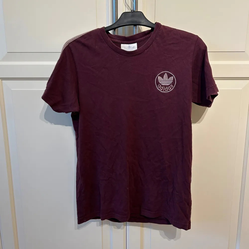 Säljer en snygg vinröd stora t-shirt  från Adidas i mycket bra skick. Den har korta ärmar och klassiska Adidas-ränder på axlarna. Perfekt för både vardag och träning. Märket syns på bröstet och materialet är mjukt och bekvämt.. T-shirts.