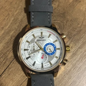 Elegant klocka med grått läderarmband - Säljer en stilren klocka från CURREN Design med ett grått läderarmband och guldiga detaljer. Urtavlan är vit med blå och röda accenter, vilket ger en modern touch. Perfekt för både vardag och fest! 🕒✨