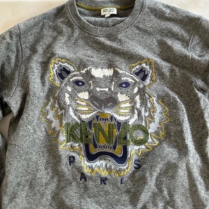 Grå sweatshirt från Kenzo - Säljer en snygg grå Kenzo sweatshirt i storlek S. Tröjan har ett coolt broderat tigerhuvud i flera färger på framsidan. Den är långärmad och perfekt för både höst och vår. Materialet känns mjukt och bekvämt, perfekt för vardagsbruk. Passar bra till jeans eller chinos för en avslappnad look.