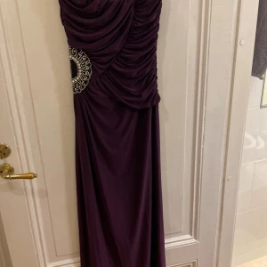 Mörklila långklänning med pärldetalj - Elegant mörklila långklänning med draperad design och vacker pärldetalj i midjan. Perfekt för festliga tillfällen. Klänningen har en smickrande passform och är tillverkad i ett mjukt, flödande material.