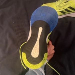 Adidas löparskor i neongrön och blå - Säljer ett par Adidas handbolls skor  i neongrön med blå detaljer. Skorna har svarta ränder på sidorna och en vit sula med blå och svarta inslag. De är i bra skick och perfekta för träning. Skorna har snörning för en säker passform.