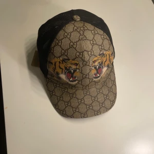 Gucci keps med tigertryck - Säljer en snygg Gucci keps med det klassiska GG-mönstret och coola tigertryck på framsidan. Kepsen har en beige och brun färgkombination med svart mesh på baksidan för extra ventilation. Perfekt för att ge din outfit en lyxig touch!